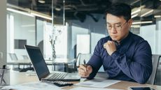 A man sits at his desk in an office and looks at paperwork in front of his open laptop.