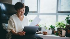 An older woman looks at paperwork with her laptop on her lap while she sits in a big chair.