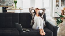 A woman puts her hands on her head and leans back on her sofa in a gesture of regret, her laptop sitting nearby.