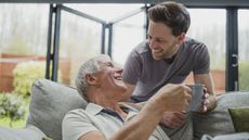 A father and son smile at each other as the son hands his father a cup of coffee.