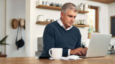 A middle-aged man works on his laptop next to paperwork sitting on his kitchen table.